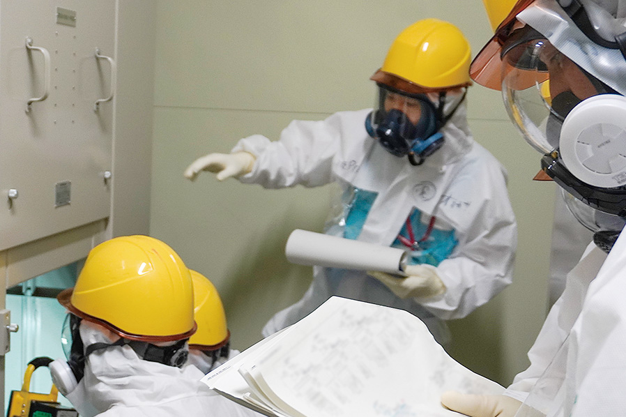4号機タービン建屋内で電路遮断の点検を行う作業員の様子(全面マスクとカバーオール装着)