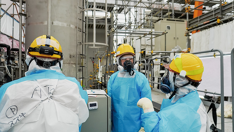 工業施設内で防護服・ヘルメット・防毒マスクを着用し、機器周辺で作業内容を確認する3人の作業員