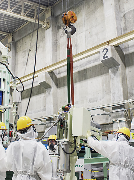 工場内で複数の作業員が遠隔・機械化設備を使用し、大型タンクの除染作業を進めている様子
